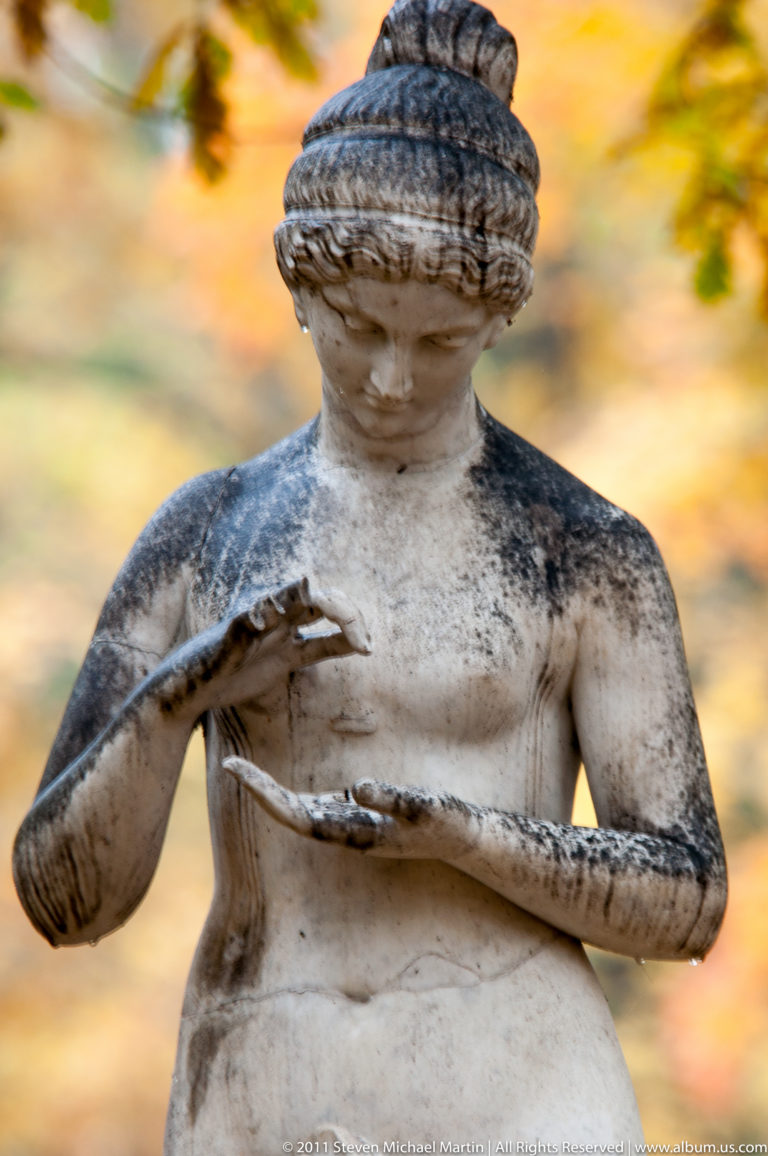 Weathered classical female statue holding a shallow object, with dark staining across the head and torso.