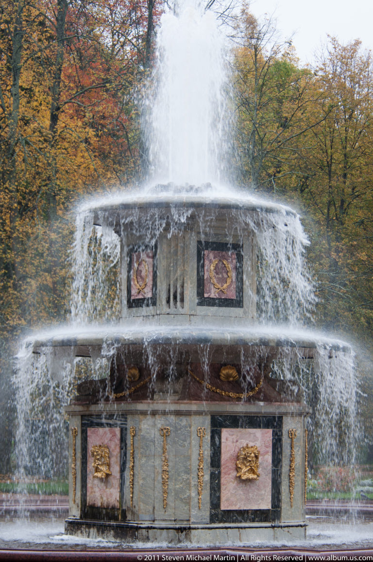 Tiered architectural fountain with a tall central jet, cascading water, colored panels, and gilded relief ornaments.