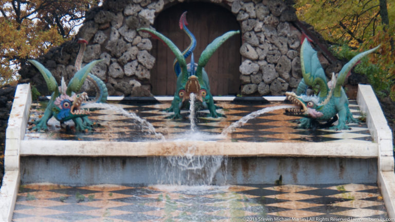 Three colorful dragon figures spouting water across the checkerboard surface before a rocky grotto entrance.