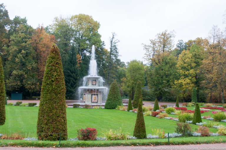 Formal lawn with flower beds and conical topiary leading to a tiered fountain against autumn trees.