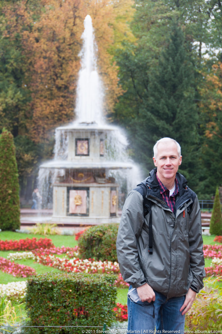 Gray-haired visitor in a dark jacket standing before a tiered fountain and colorful formal flower beds.