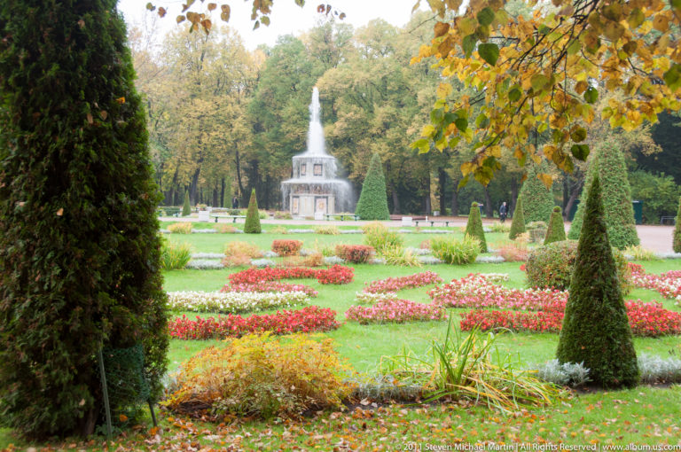 Tiered fountain seen across flower beds and conical topiary, framed by yellow autumn leaves and trees.