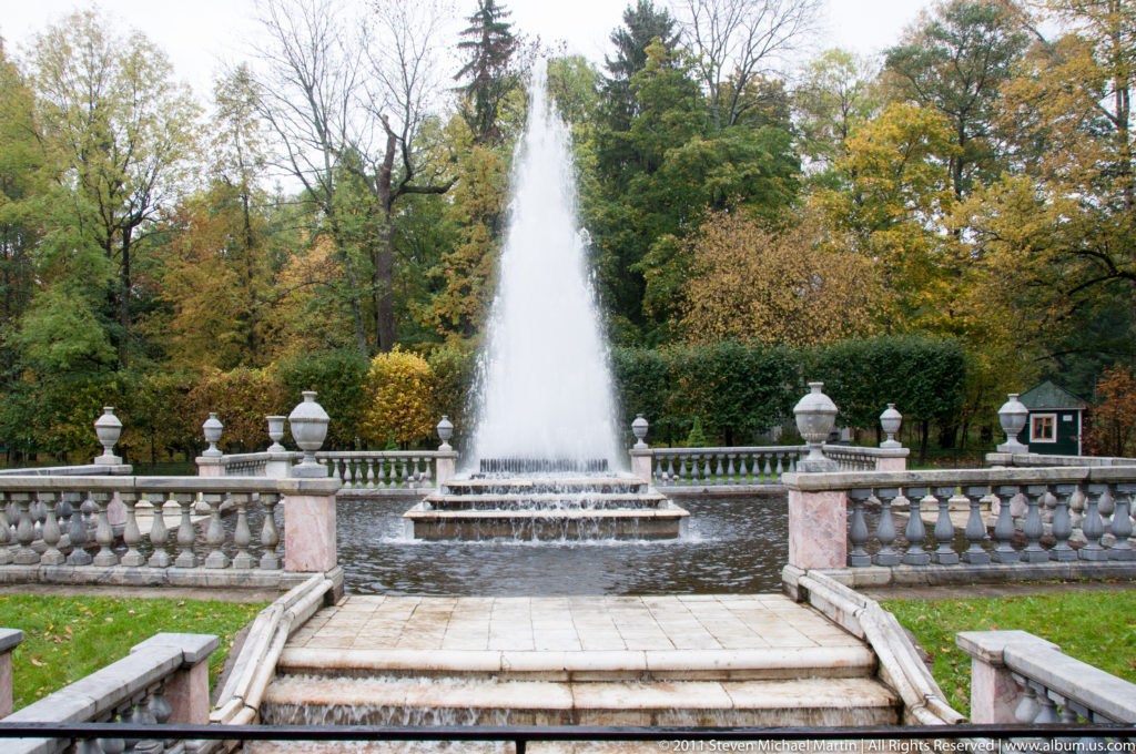 Tall fountain jet in a circular basin framed by stone balustrades, steps, and urn-topped posts.