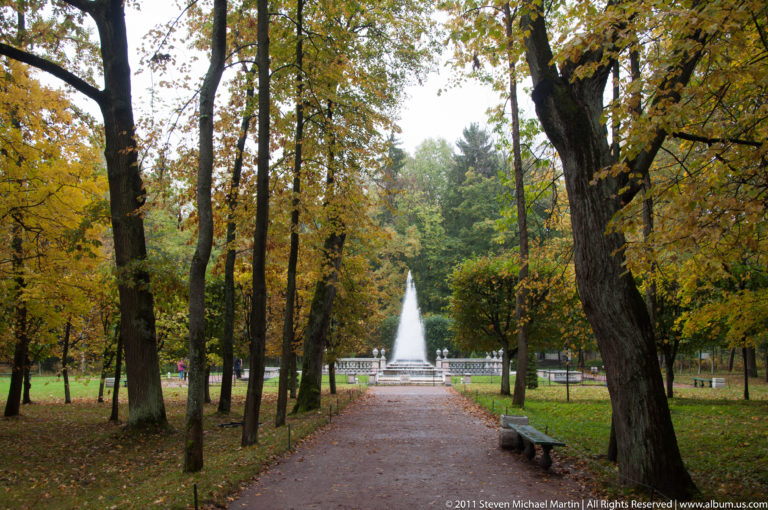 Gravel path through autumn woodland leading straight toward a tall fountain jet in the distance.