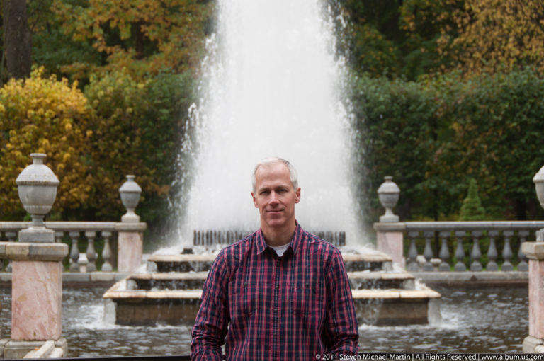 Gray-haired visitor in a red plaid shirt standing before a tall fountain jet and stone balustrade.
