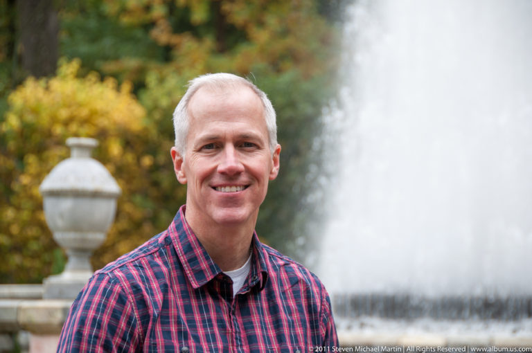 Gray-haired visitor in red plaid beside a tall fountain jet, with an urn-topped stone post at left.