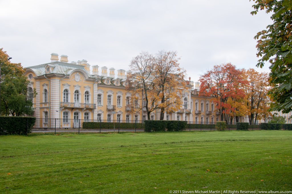 Long pale-yellow Baroque palace façade with white ornament beside a broad lawn and autumn trees.