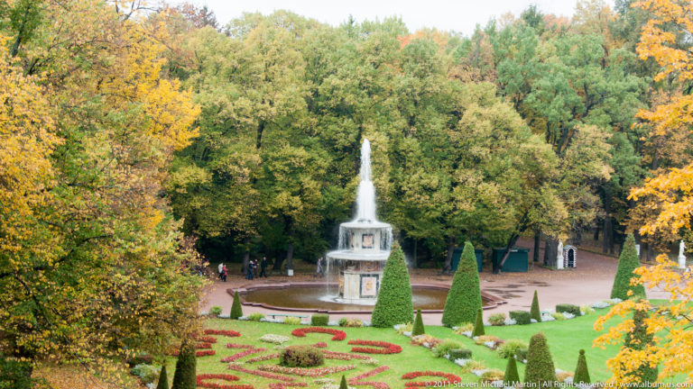 Elevated view over patterned flower beds and conical topiary toward a tiered fountain and autumn woods.