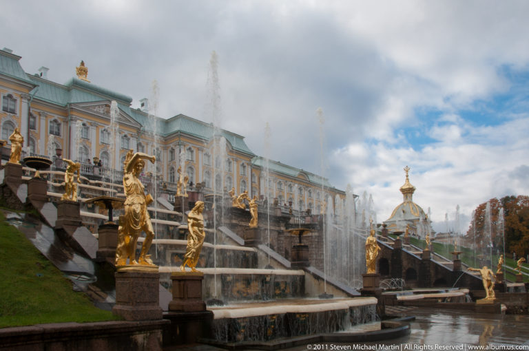 Side view of Grand Cascade terraces, gilded statues, fountain spray, and the Grand Palace under cloudy sky.
