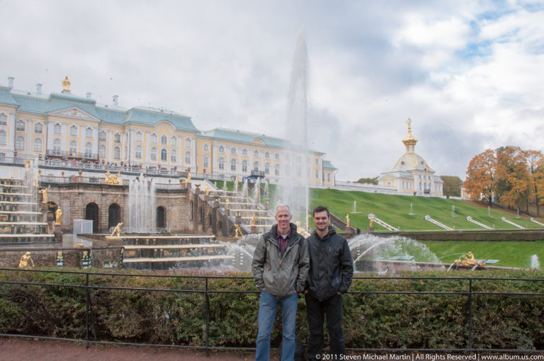 Two men at a railing with Peterhof Grand Cascade, fountains, lawns, and Grand Palace behind.