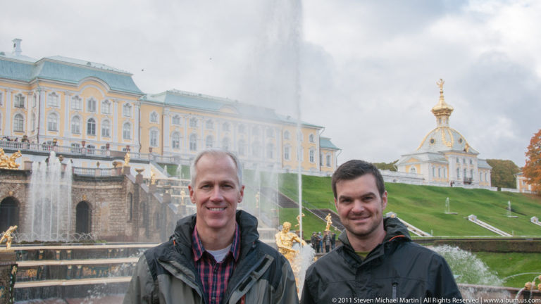 Two men standing together before the Grand Cascade and long yellow-and-white Grand Palace façade.