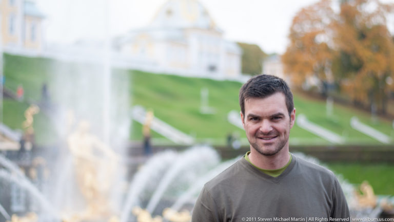 Smiling man standing before blurred Peterhof fountains, gilded sculpture, green slope, and palace buildings.