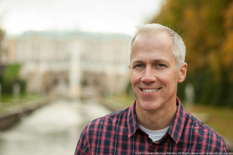 Smiling man in a plaid shirt with Peterhof palace gardens and water blurred behind him.