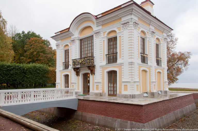 Small yellow-and-white pavilion with tall windows, iron balcony, brick base, and narrow water channel.