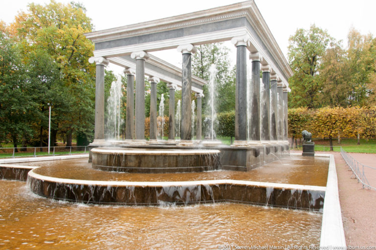 Classical colonnade above tiered fountain basins with water spilling over curved edges in an autumn park.