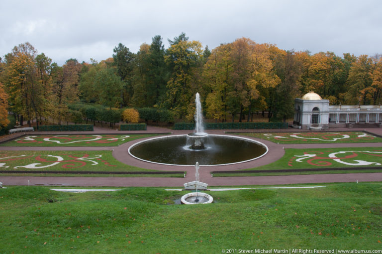 Symmetrical patterned parterre around a circular fountain, with autumn trees and a small pavilion beyond.