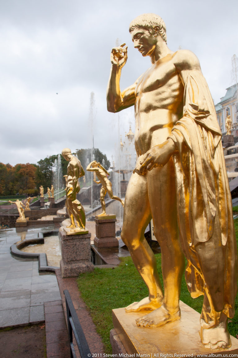 Large gilded male statue in foreground with smaller gilded fountain figures and water jets behind.