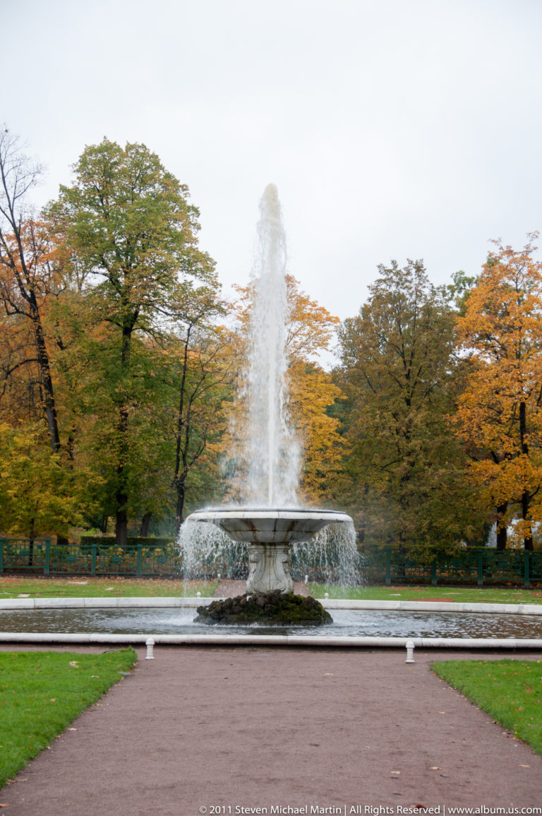 Circular garden fountain centered on a path and lawn, backed by autumn trees.