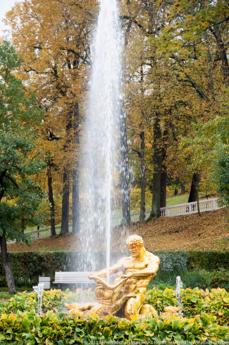 Tall fountain jet rising behind a reclining gilded figure amid green plants and autumn trees.
