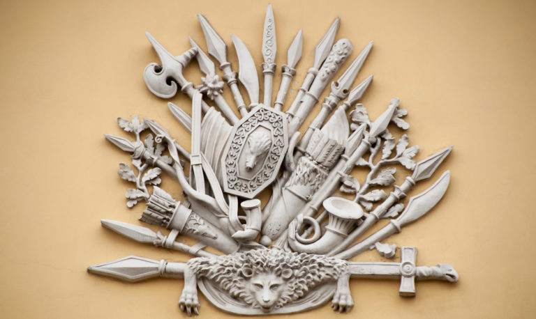 White sculptural relief of spears, banners, shield, helmet, trumpet, axes, and lion mask on an ocher wall.