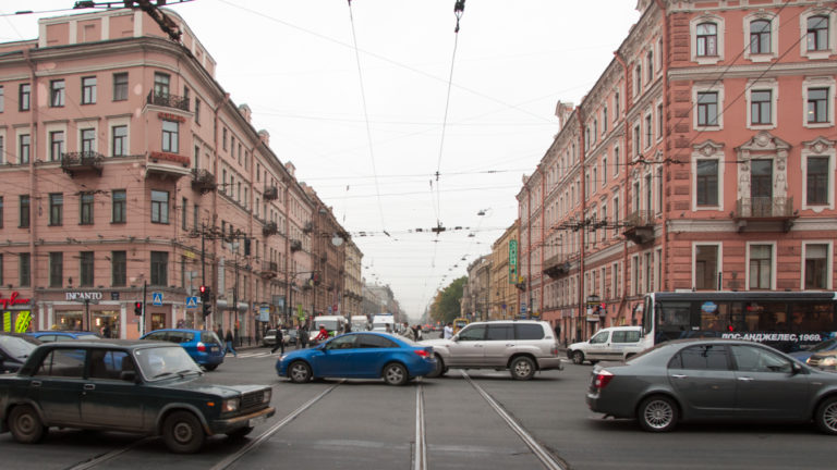 Busy St. Petersburg avenue with cars, paired rails, overhead wires, and continuous rows of historic multistory buildings.