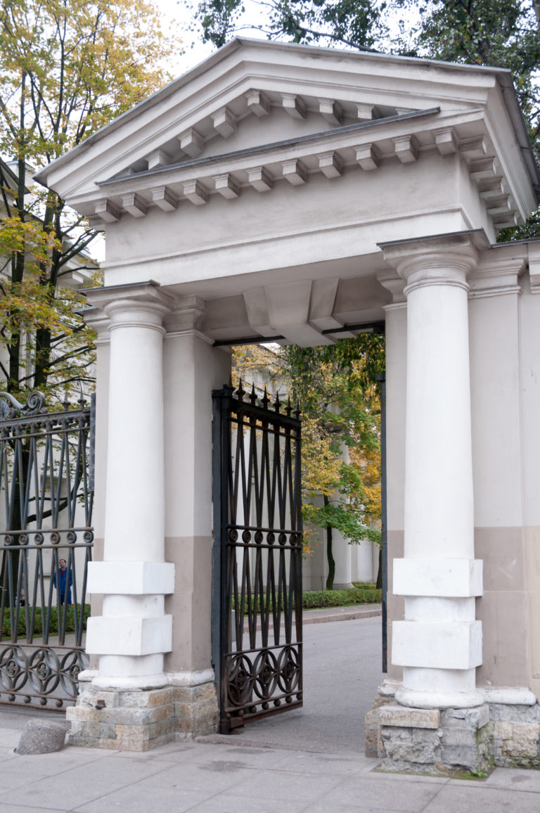 White Doric gateway with paired columns and open black iron gates beneath autumn trees.