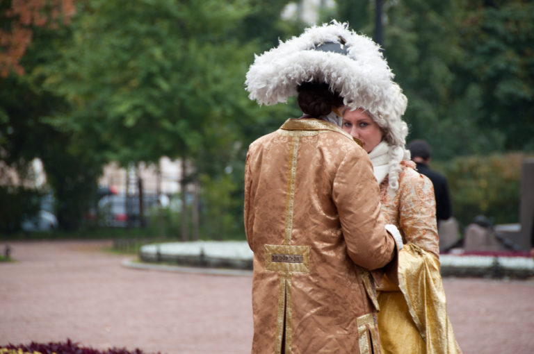 Two people in elaborate gold and white eighteenth-century-style costumes standing together in a leafy city park.