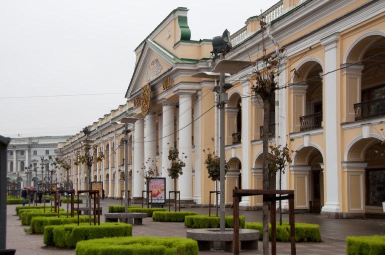 Ocher-and-white Great Gostiny Dvor façade with columns, arched gallery openings, shops, small trees, and clipped hedges.