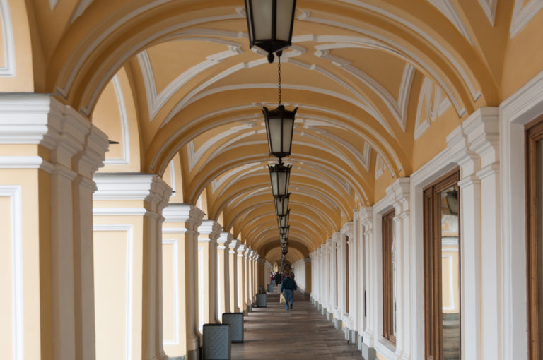 Long ocher-and-white vaulted arcade with repeated piers, shop windows, hanging lanterns, and pedestrians.