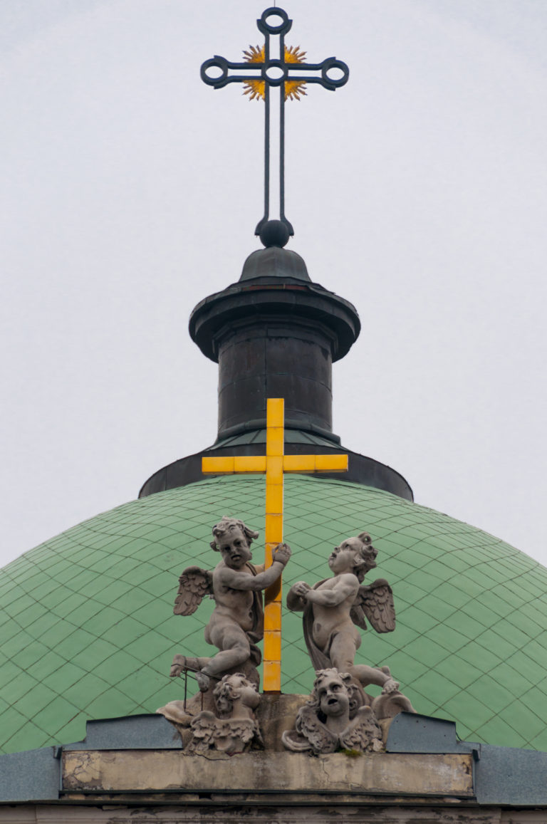 Green tiled dome with stone figures, dark cupola, cross, and gilded ornament against a gray sky.