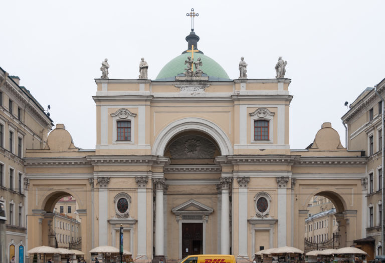 Pale classical church façade with large central arch, side passages, rooftop statues, green dome, and cross.
