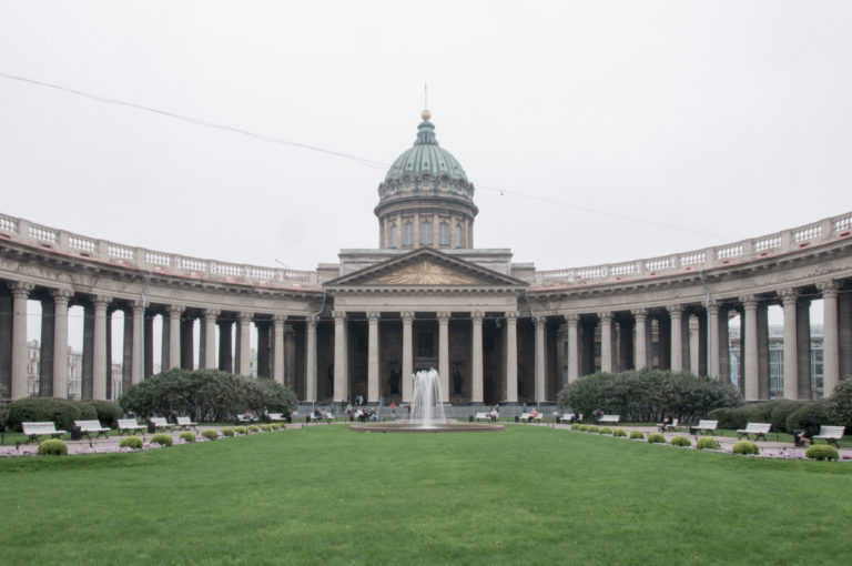 Wide symmetrical view of Kazan Cathedral with green dome, central portico, curving colonnade, lawn, and fountain.