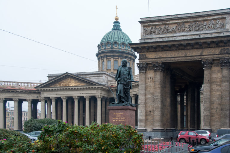 Bronze military monument on a red-granite pedestal before the colonnade and green dome of Kazan Cathedral.