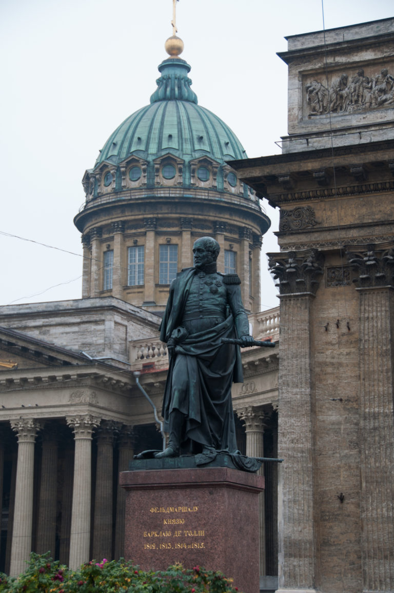 Rear view of the bronze Mikhail Kutuzov monument on a red-granite pedestal with Kazan Cathedral and green dome behind.