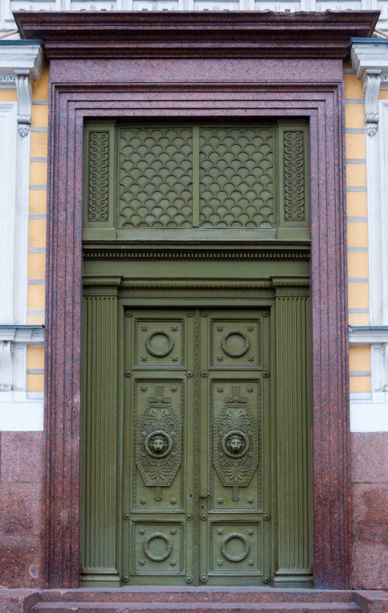 Large green double door with relief medallions and patterned transom framed by polished red granite.