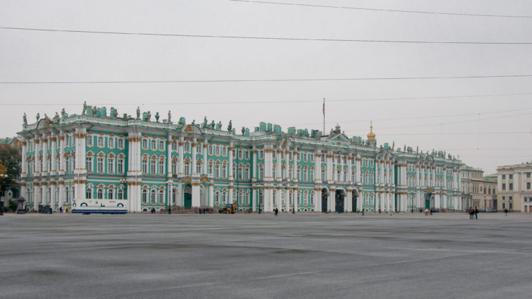 Long oblique view of the Winter Palace across a broad, mostly empty Palace Square beneath overhead wires.
