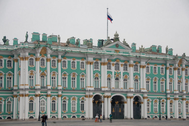 Green-and-white Winter Palace façade with three central gates and small pedestrians crossing Palace Square.