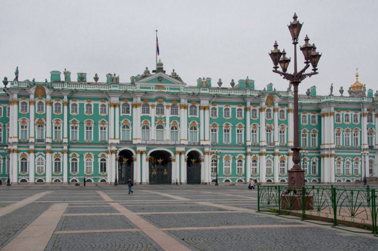 Winter Palace across patterned Palace Square paving, with an ornate streetlight in the foreground and Russian flag above.