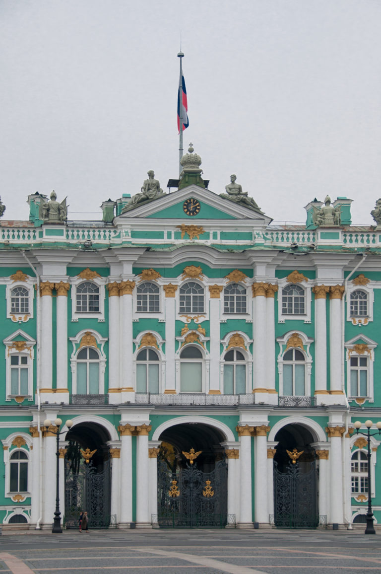 Central Winter Palace façade with three arched gates, white columns, gilded ornament, roof statues, and Russian flag.