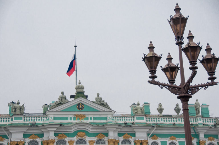Ornate multi-lantern streetlight beside the Russian flag and sculpted roofline of the Winter Palace.
