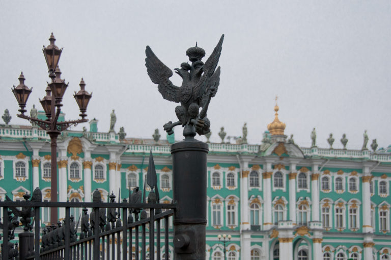 Dark double-headed eagle atop an iron post, with the green-and-white Winter Palace softly focused behind.