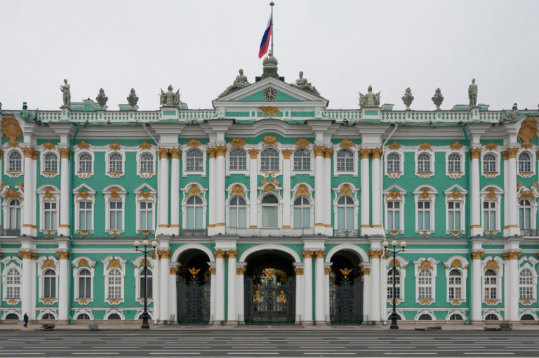 Broad frontal view of the green-and-white Winter Palace façade with three arched gates, roof statues, and Russian flag.