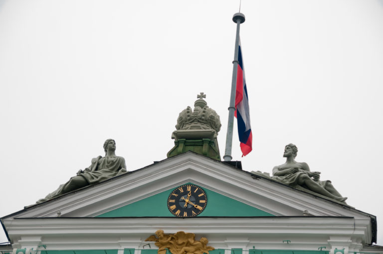 Russian flag above the Winter Palace pediment with clock, crowned crest, and seated roofline statues.