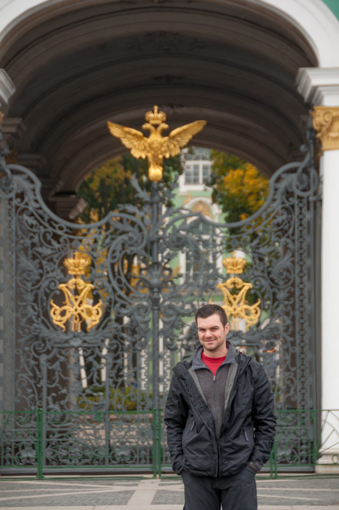 Man standing before an ornate dark iron Winter Palace gate decorated with large gilded imperial eagles and monograms.