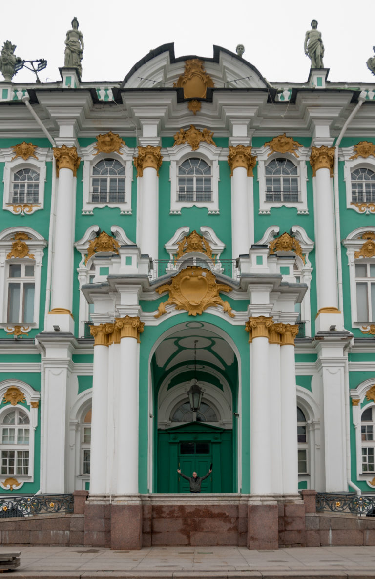 Frontal view of the Winter Palace central gateway with white columns, gilded Baroque ornament, and green walls.