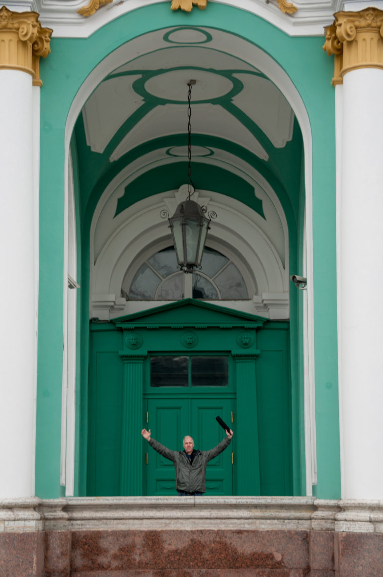 Small man with arms outstretched beneath a tall Winter Palace arch with green doors and hanging lantern.