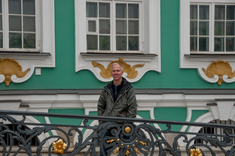 Man standing behind an ornate black-and-gold railing against the green-and-white Winter Palace façade.