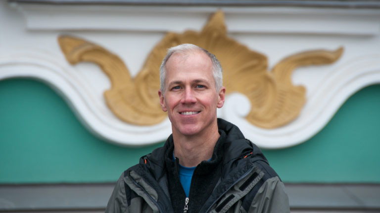 Smiling man in a dark jacket posed before the blurred green, white, and gilded Winter Palace façade.