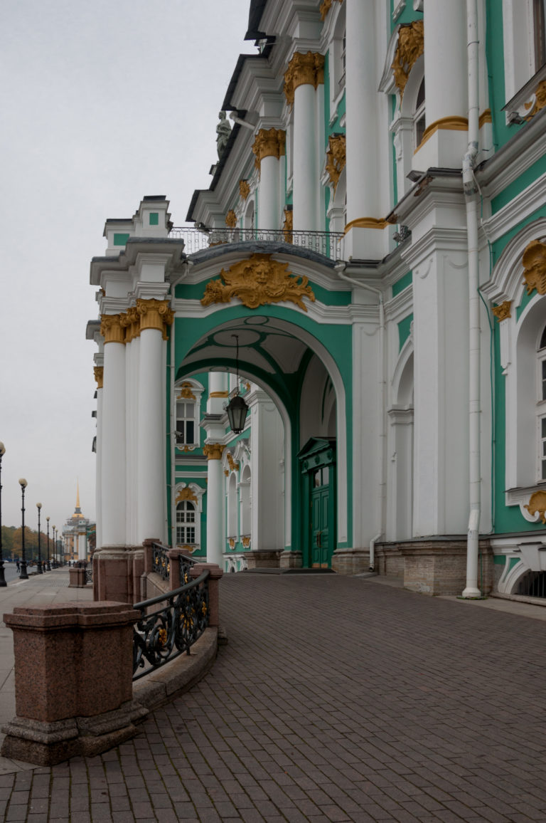 Oblique view of a Winter Palace arched gateway with white columns, gilded capitals, lantern, and green façade beyond.