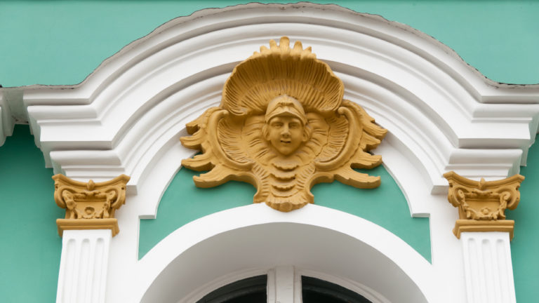 Gilded female face and shell ornament above a white arched window surround on the green Winter Palace façade.